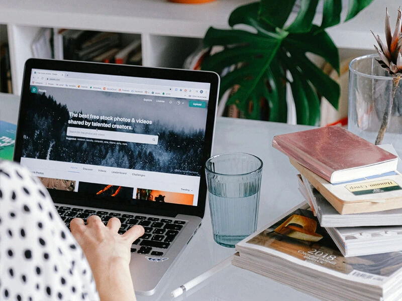 Online Research and Study A person's hands are shown typing on a laptop displaying a stock photo website. A glass of water and a stack of books are on the white desk next to the computer